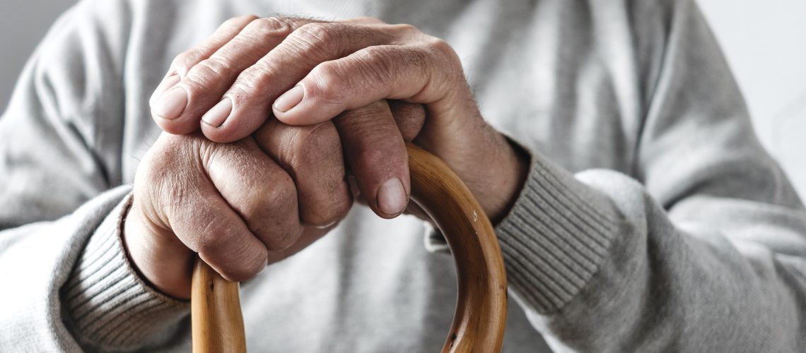 Close up details of the folded hands of an elderly man resting on a walking cane in a mobility and health concept